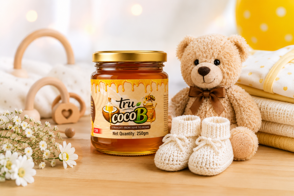 Tru-CocoB honey jar with baby gift items including teddy bear, knitted booties, folded baby clothes, and flowers